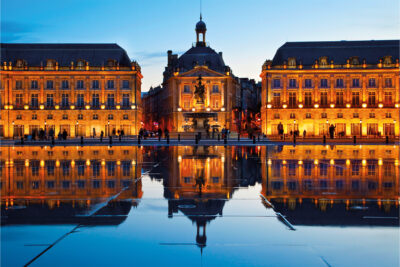 Buildings lit up with lights in, you can see their reflection on the floor on the wet surface