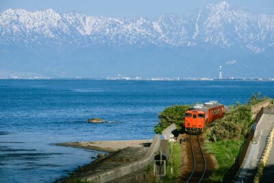 Red Local Train Passing by Toyama Bay with Tateyama Mountain Range in Background – Toyama, Japan, May 8, 2025